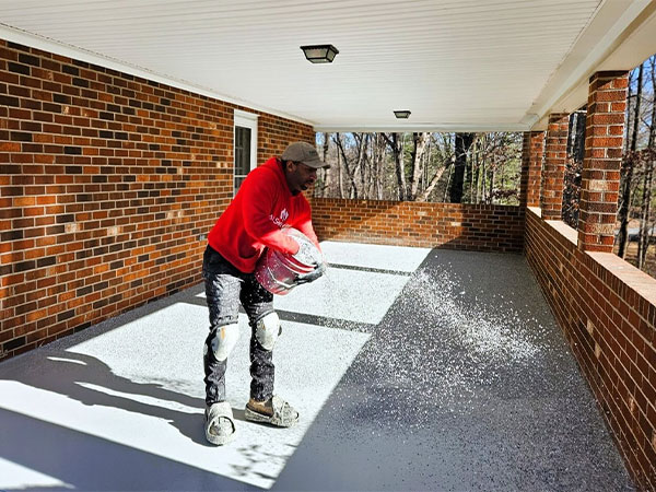 Man applying outdoor concrete coatings to a brick porch. He's wearing knee pads and a red sweatshirt, spreading decorative flakes over the fresh coating.